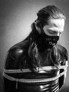 Une personne au cheveux long en latex avec un masque en latex est attaché par des cordes de shibari