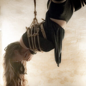 Une jeune fille est suspendu en l'air par des cordes de shibari, sont masque de voleuse est retombé et elle le tient dans la bouche.
